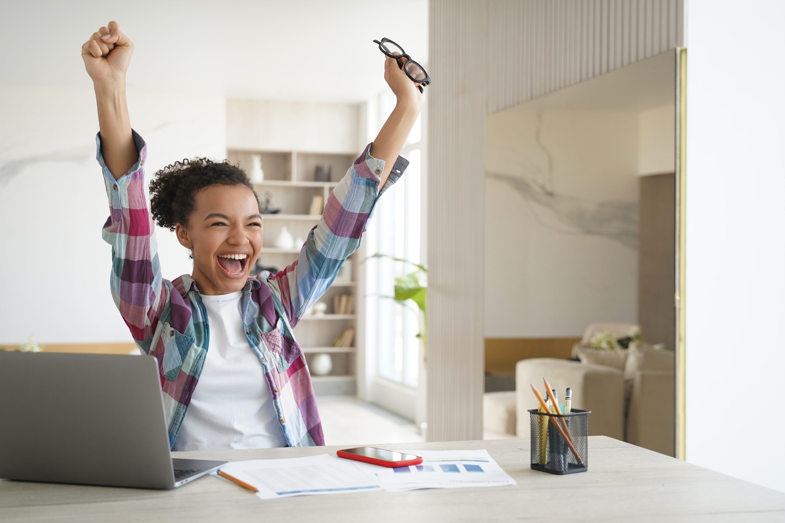 Excited student cheering with arms raised at her desk after seeing test results on her laptop, representing the confidence and success Tutor of Dragons' SAT/ACT test prep tutoring helps students achieve.