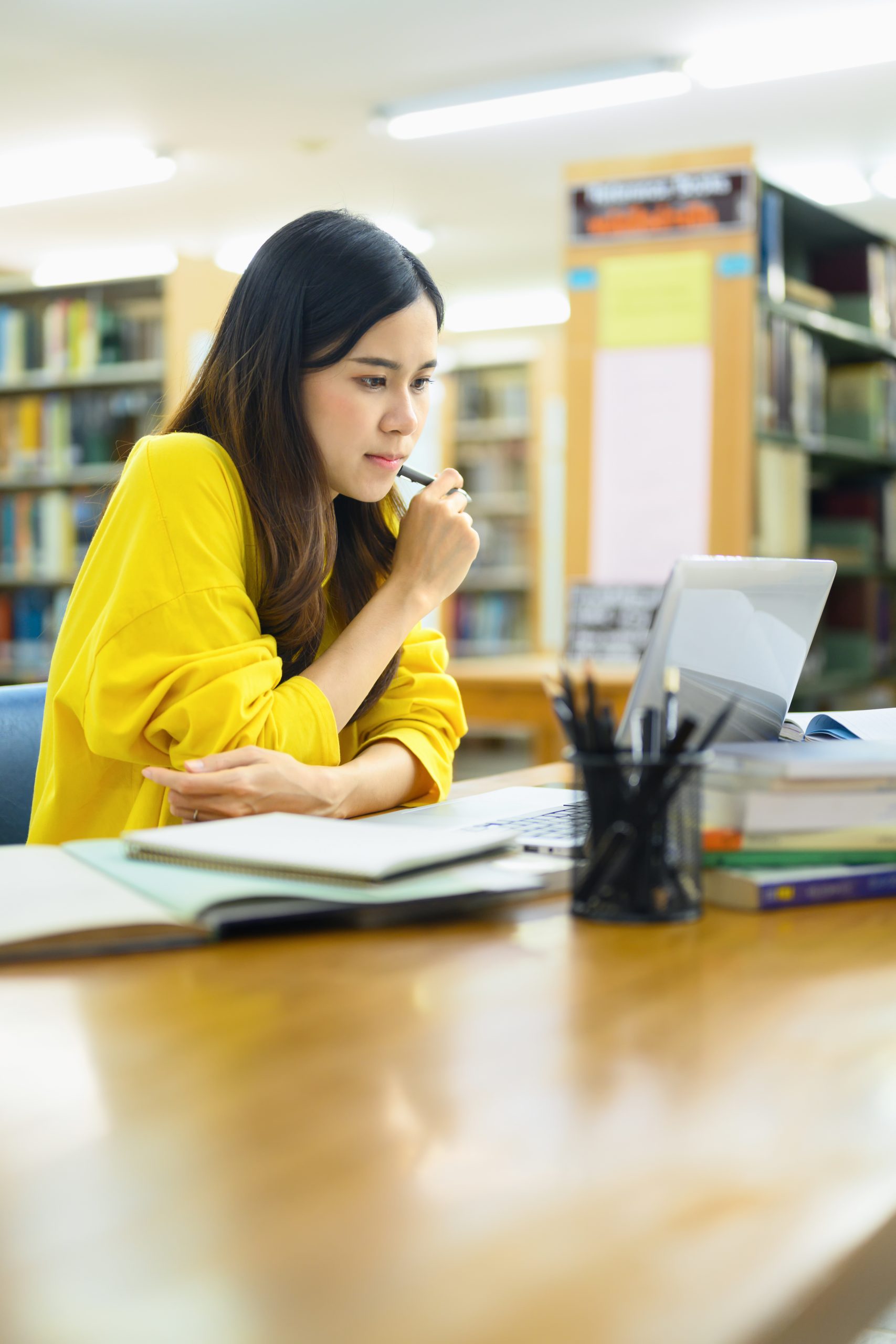 Student in a library thoughtfully reviewing study materials on a laptop, surrounded by notebooks and textbooks, reflecting the focused, strategic approach Tutor of Dragons brings to SAT/ACT test prep tutoring in Southlake, Texas.